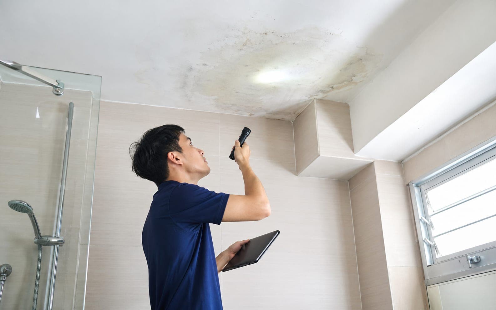 Contractor shining a torch on a water-stained bathroom ceiling while inspecting a leak in a Singapore flat
